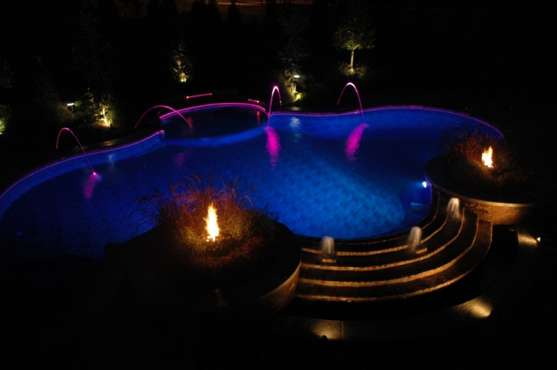 Multi-color pool lights, water features, and fire features make this pool by Burton Pools & Spas, your local inground pool builder near me, a spectacular display at night.